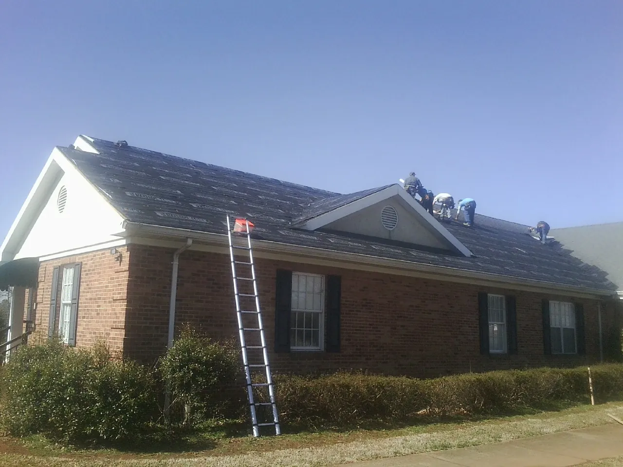 Crew tearing shingles off a brick ranch roof with a ladder set — Metal Roof Repair work by our crew in Intervale