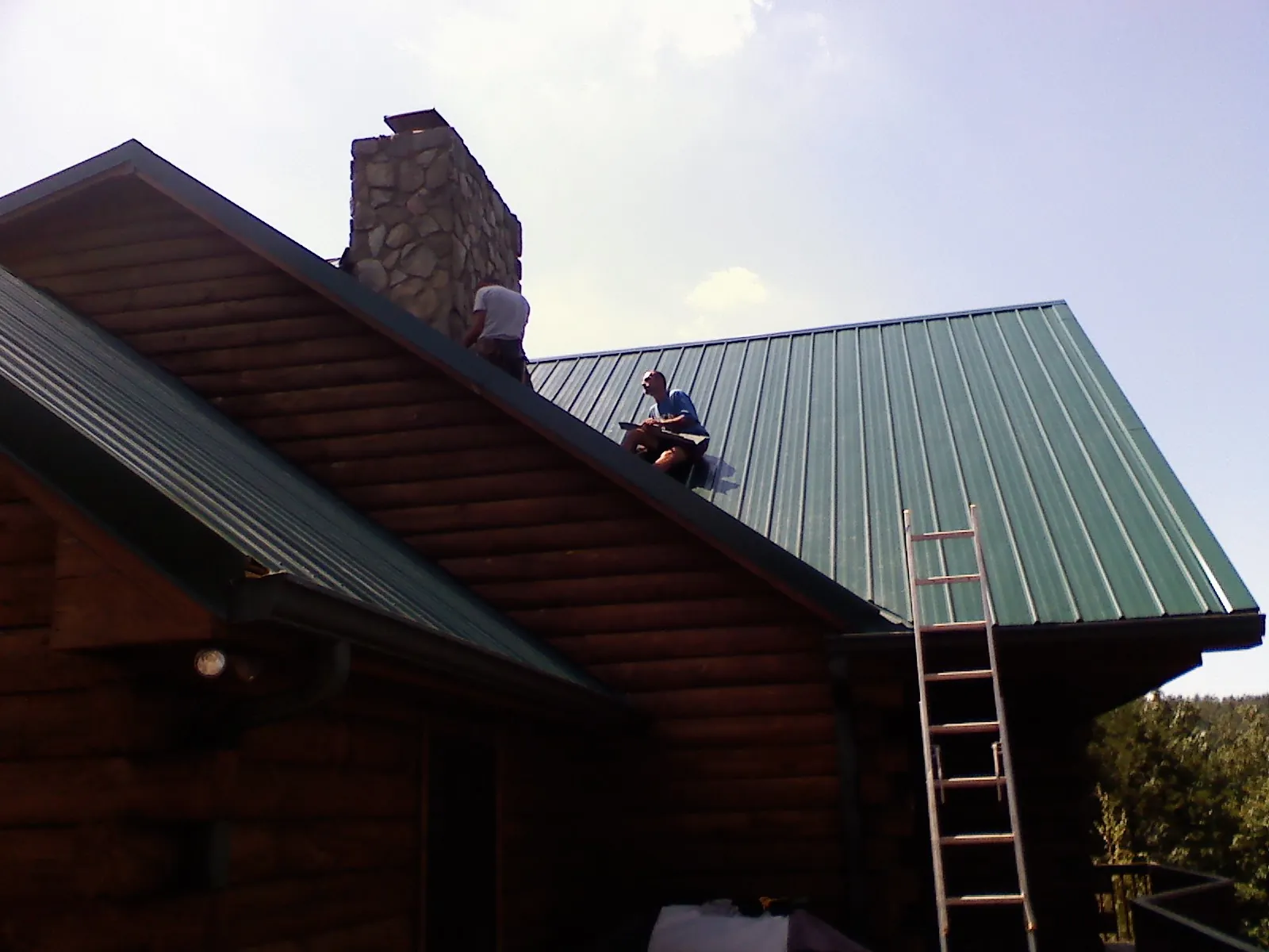 Roofers installing green metal panels beside a stone chimney on a log home in Intervale